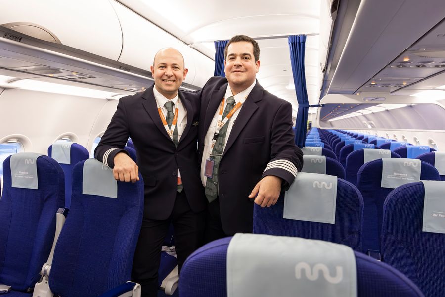 The image shows two airline pilots standing together inside the cabin of a commercial airplane. Both are dressed in formal pilot uniforms with jackets, ties, and lanyards, and are smiling at the camera with one arm around each other. The cabin features rows of blue seats with light grey antimacassars displaying a logo. The setting is well-lit and clean, suggesting a professional and welcoming atmosphere onboard.